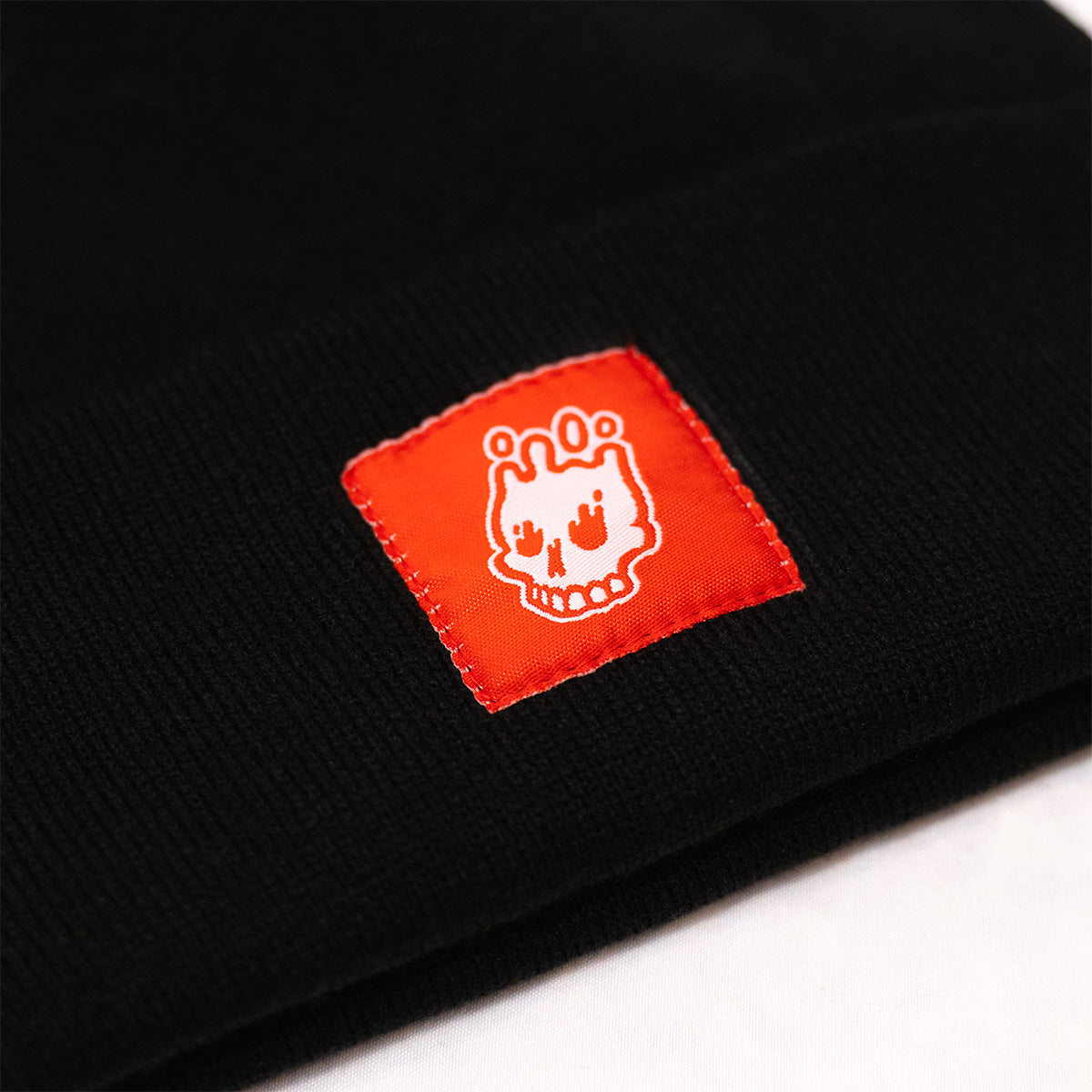 Close up of black cuff beanie with white Rubber Bones Icon logo on a red patch.