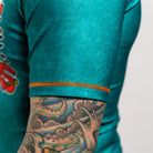 Close-up of the opposite sleeve of the same teal rash guard, showing orange stitching detail and a tattooed forearm. This sleeve does not have a patch or logo.