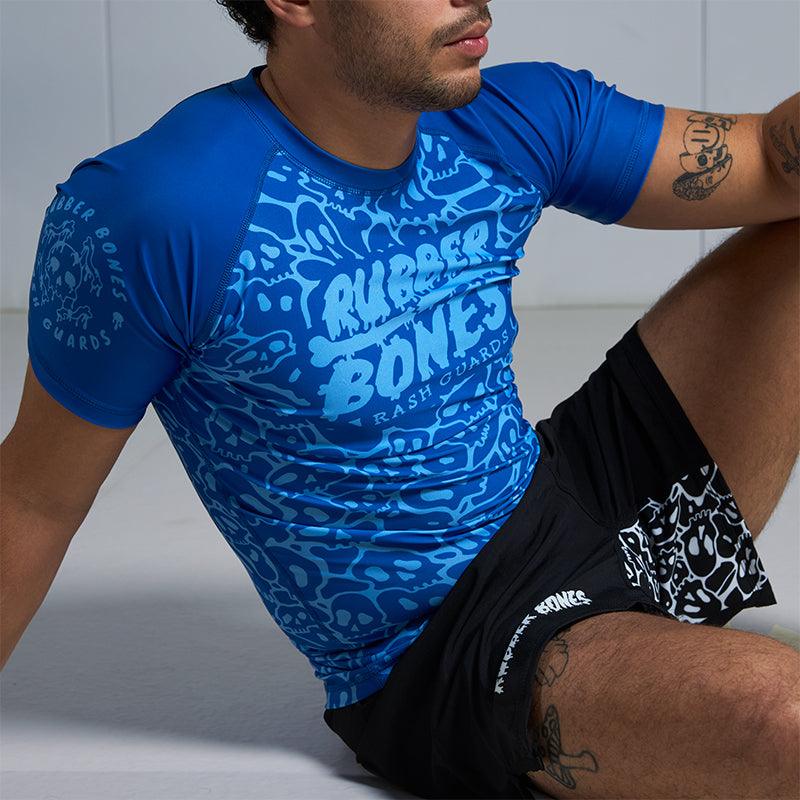 On-body seated view of Blue Ranked Rash Guard paired with Rubber Bones black competition grappling shorts.