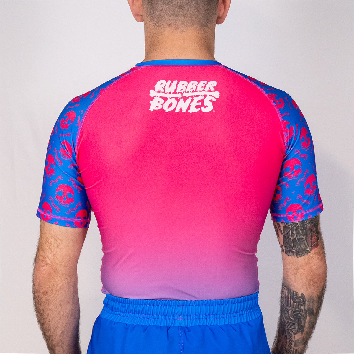 Back view of the Rubber Bones Toxic Berry men’s grappling rash guard with bright pink fade body, blue sleeves, and skull artwork pattern.