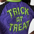 “Trick or Treat” text in dripping green font with eyeball graphic on back of purple women’s rash guard by Rubber Bones.
