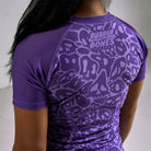 On-body 3/4 back close-up of Women's Purple Ranked Rash Guard highlighting the Rubber Bones logo and skull web design.