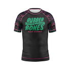 Kids' BJJ rash guard featuring a bold 'Rubber Bones Rash Guards' logo in dripping green text on a black body. The short sleeves showcase a neon-inspired pattern of pink, green, and purple drippy smiley faces. Finished with pink stitching, this rash guard combines durability, comfort, and standout style for young grapplers.