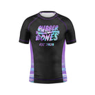 A black kids short-sleeve BJJ rash guard with "Rubber Bones" in a holographic-style font, blending purple, blue, and pink hues. "Est. 2020" is written below in a stylized font. The side panels feature a gradient holographic effect.