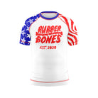 A kids short-sleeve BJJ rash guard with a white base and a distressed red, white, and blue design. The front features "Rubber Bones" in red with a grungy, weathered texture, along with "EST. 2020" beneath it. The sleeves are decorated with USA flag elements—red and white waves on one side and a blue field with white stars on the other.