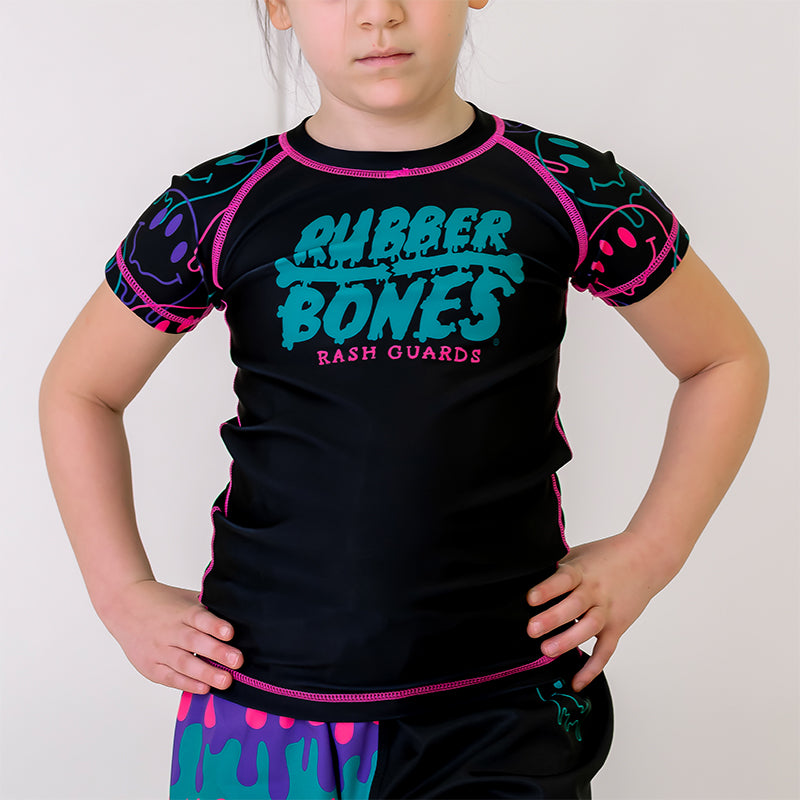 Bubble Gum Kids BJJ Rash Guard, front view, featuring a black body with a green and pink Rubber Bones logo and purple, green, and pink dripping alien smiley faces on the sleeves. Pink Stitching.