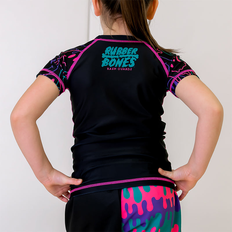 ubble Gum Kids BJJ Rash Guard, back view, showcasing a black body with a green and pink Rubber Bones logo at the nape of the neck and dripping alien smiley faces-patterned sleeves. Pink Stitching.