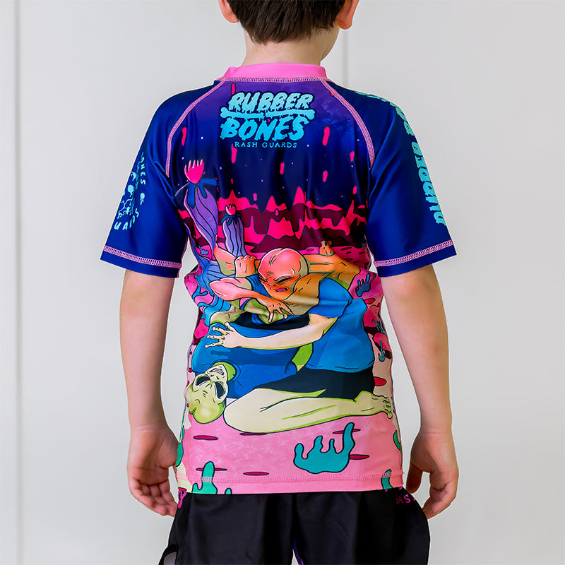 Kids BJJ Invasion Rash Guard, back view, showcasing a fully illustrated alien scene in bright colors with pink stitching.