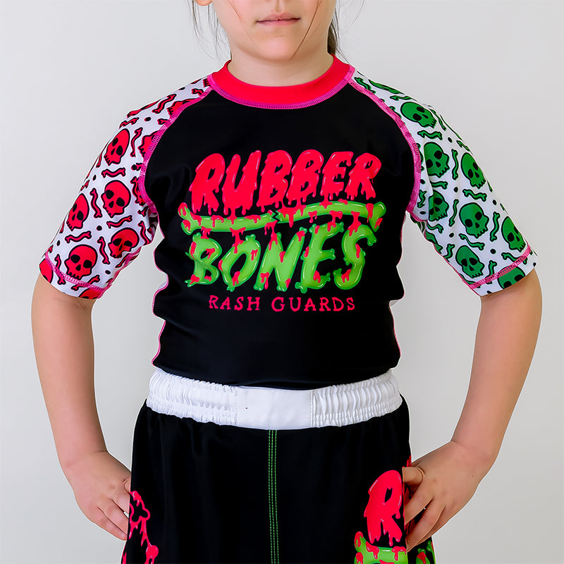 OG Kids BJJ Rash Guard, front view, featuring a black body with pink and green Rubber Bones logo, white sleeves, green skull pattern on the left sleeve, and pink skull pattern on the right sleeve.