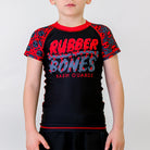 Black OG 2.0 Kids BJJ Rash Guard, front view, featuring the Rubber Bones logo on the center chest, red and grey skull pattern sleeves, and red and grey accents.