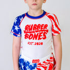 Independence Kids BJJ Rash Guard, front view, featuring a white body with a red Rubber Bones logo, trippy red stripes on the left sleeve, and white distressed stars on the right blue sleeve.