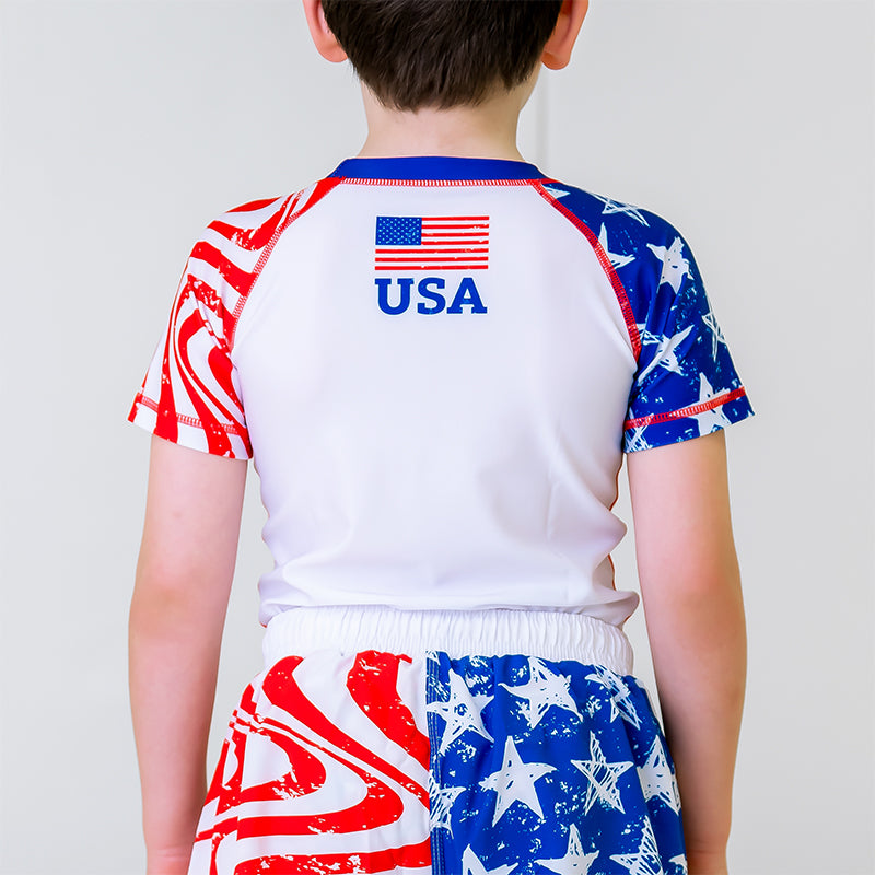 Independence Kids BJJ Rash Guard, back view, featuring a white body with an American flag logo and "USA" at the nape of the neck.