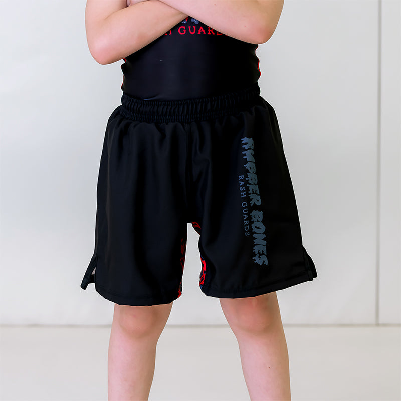 Kids BJJ OG 2.0 Shorts, front view, featuring black shorts with a grey Rubber Bones logo on the left leg, red stretch panel with grey skull pattern, and black right leg.