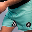 Close-up of the left side of mint green BJJ shorts featuring a black circular patch with a white Rubber Bones skull logo. A black and white checker pattern is visible in the side slit.