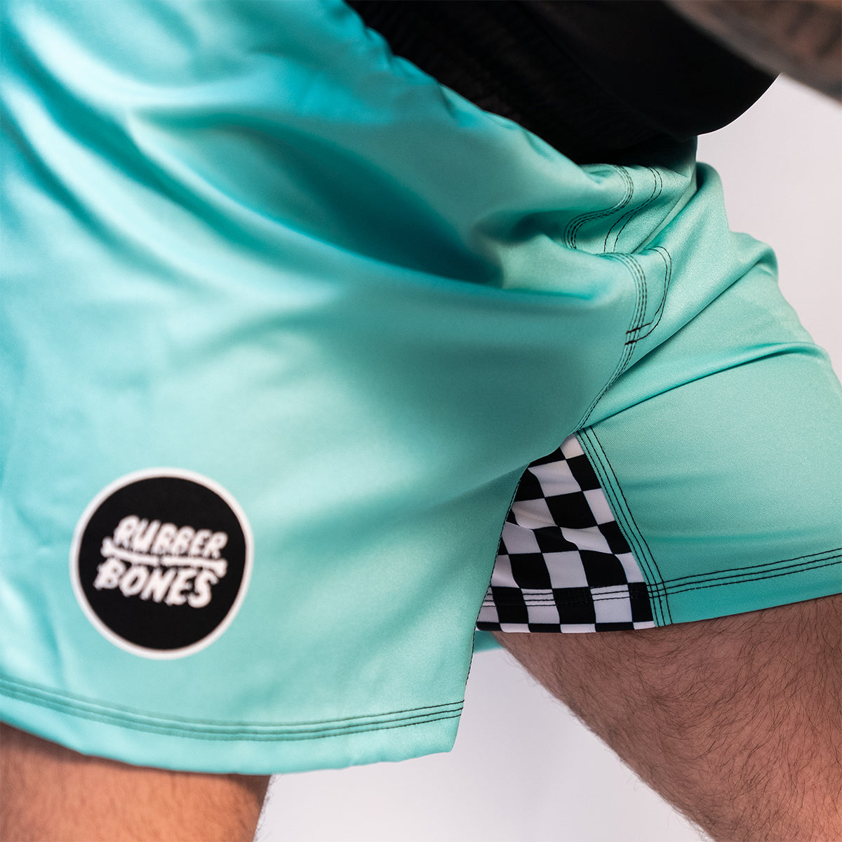 Close-up of the right side of mint green BJJ shorts showing a black circular Rubber Bones logo patch and checker detail in the side slit.