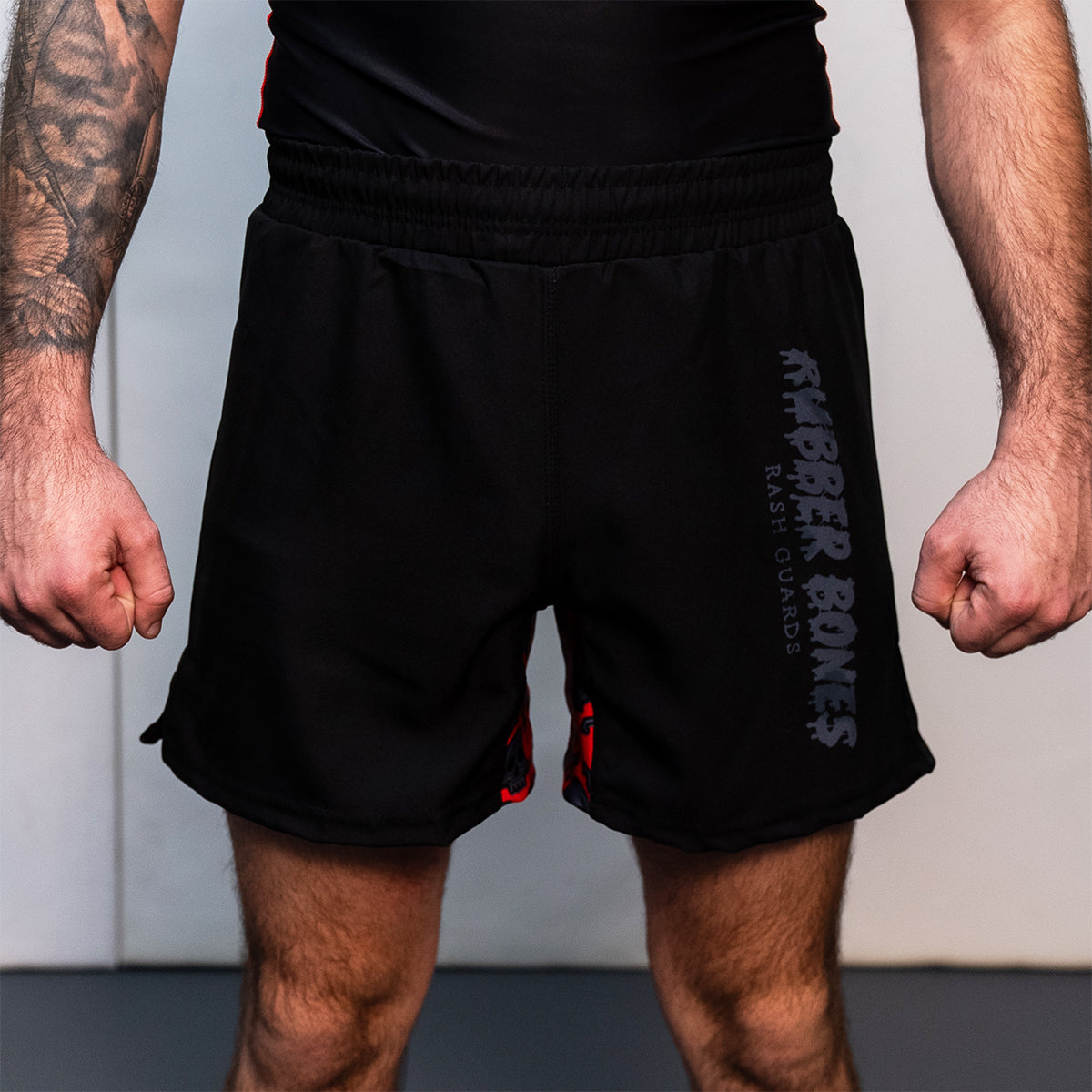 Black BJJ fight shorts with an elastic waistband and side slits. "Rubber Bones Rash Guards" logo in dark gray on the left leg. Red and black skull pattern visible on inner leg panel.