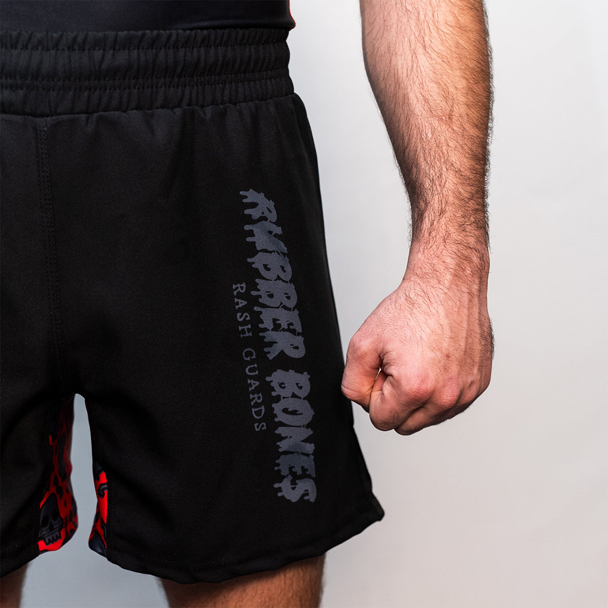 Black BJJ shorts with side slits and an elastic waistband. "Rubber Bones Rash Guards" logo in dark gray on the left leg. Subtle red skull pattern visible on inner leg panel.