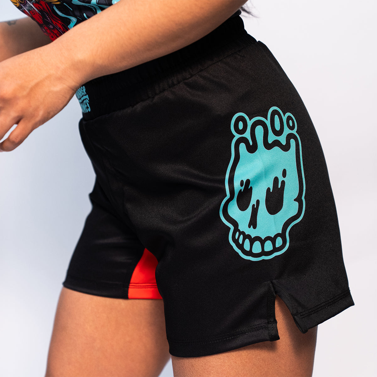 Side view of Rubber Bones Tora Oni BJJ Shorts – Black no-gi shorts with a teal melting skull graphic and a red stretch gusset.