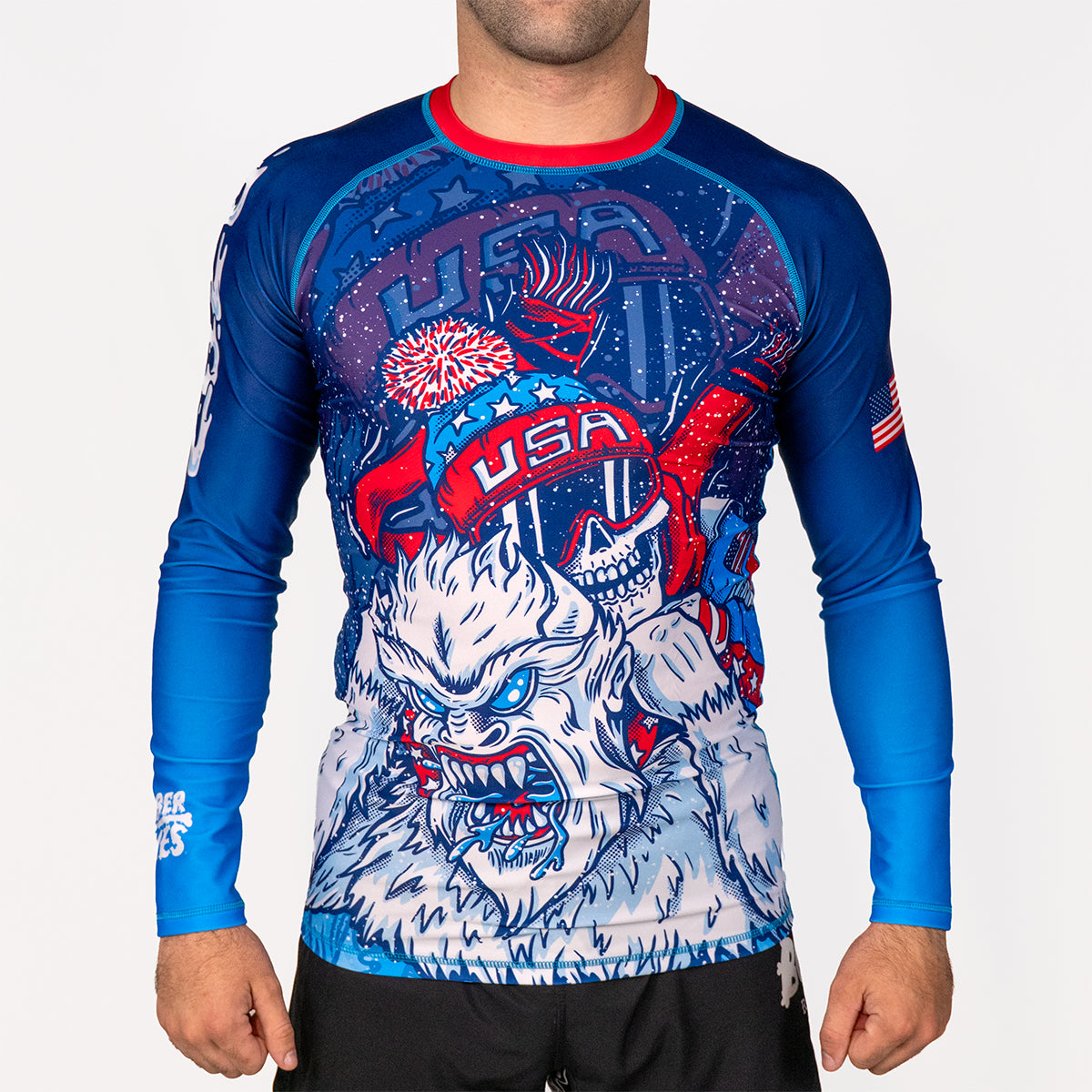 Men's Yeti Games long sleeve rash guard front view featuring skeleton skier choking Yeti artwork in patriotic red, white, and blue, full-length compression sleeves, USA flag patch, red neckline band.