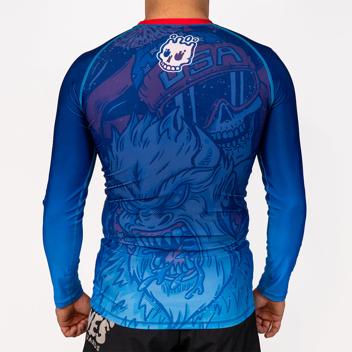 Back view of Men's Yeti Games long sleeve rash guard showing white Rubber Bones skull logo at top, skeleton skier Yeti design, red neckline band, full-length blue compression sleeves.