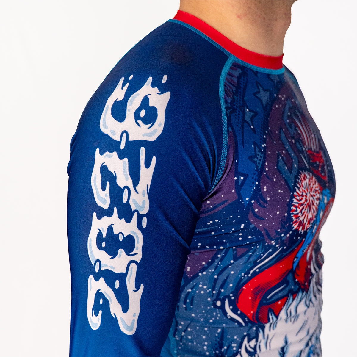 Side view of Men's Yeti Games long sleeve rash guard showing white 2026 lettering on blue sleeve, skeleton skier Yeti artwork on chest with patriotic red and blue coloring.
