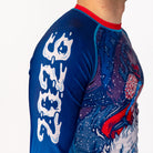 Side view of Men's Yeti Games long sleeve rash guard showing white 2026 lettering on blue sleeve, skeleton skier Yeti artwork on chest with patriotic red and blue coloring.