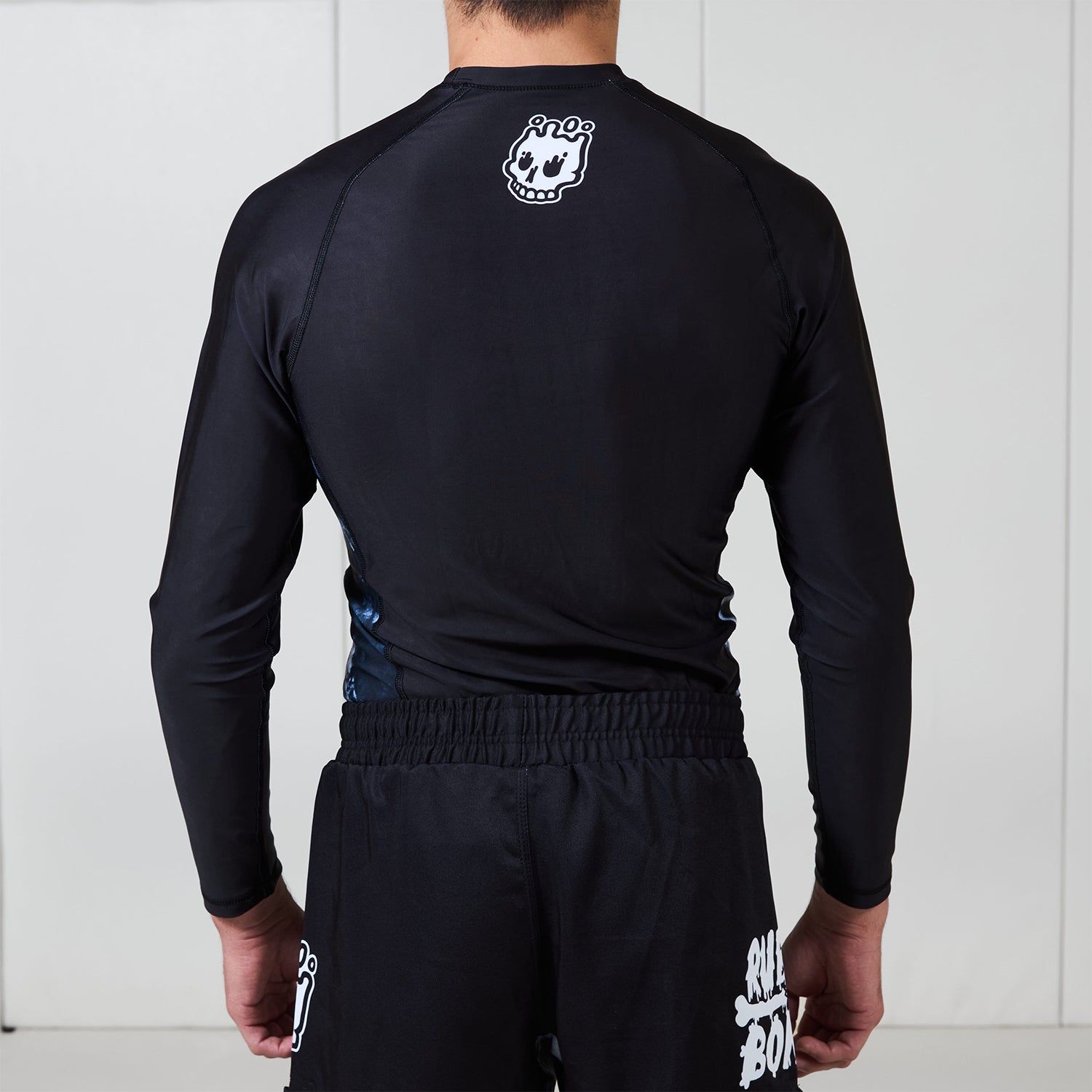 Back view of the Men’s Long Sleeve Ash BJJ Rash Guard showing black design with white melting skull Rubber Bones logo near neckline.