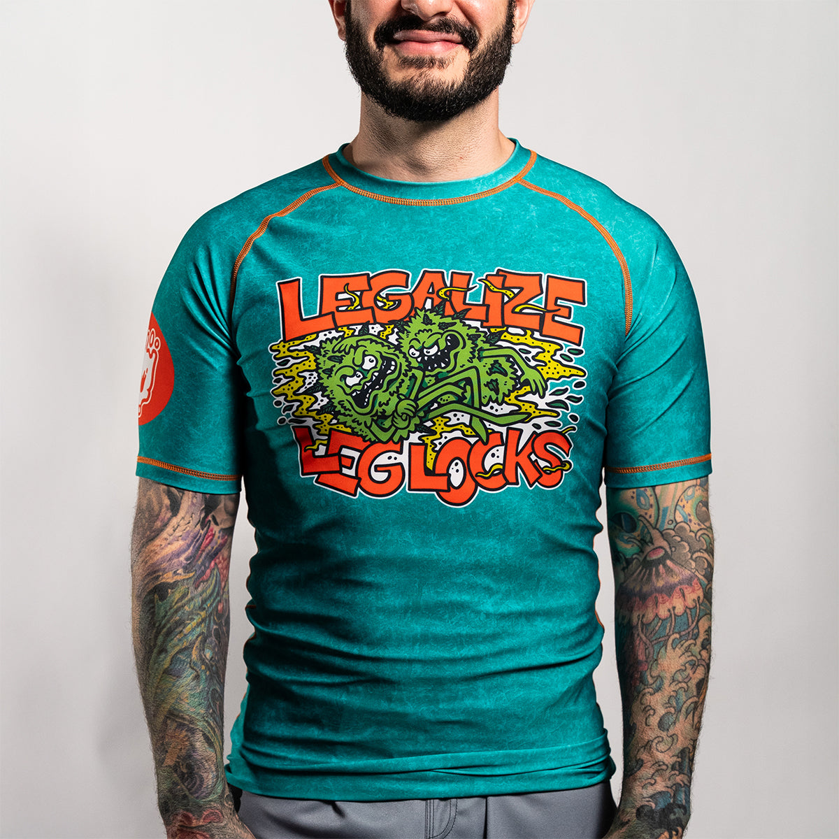 Front view of a person wearing the rash guard. The center chest features a large graphic with green cartoon creatures and the words “Legalize Leg Locks” in bold orange letters. The skull icon is visible on the right sleeve.