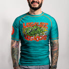 Front view of a person wearing the rash guard. The center chest features a large graphic with green cartoon creatures and the words “Legalize Leg Locks” in bold orange letters. The skull icon is visible on the right sleeve.