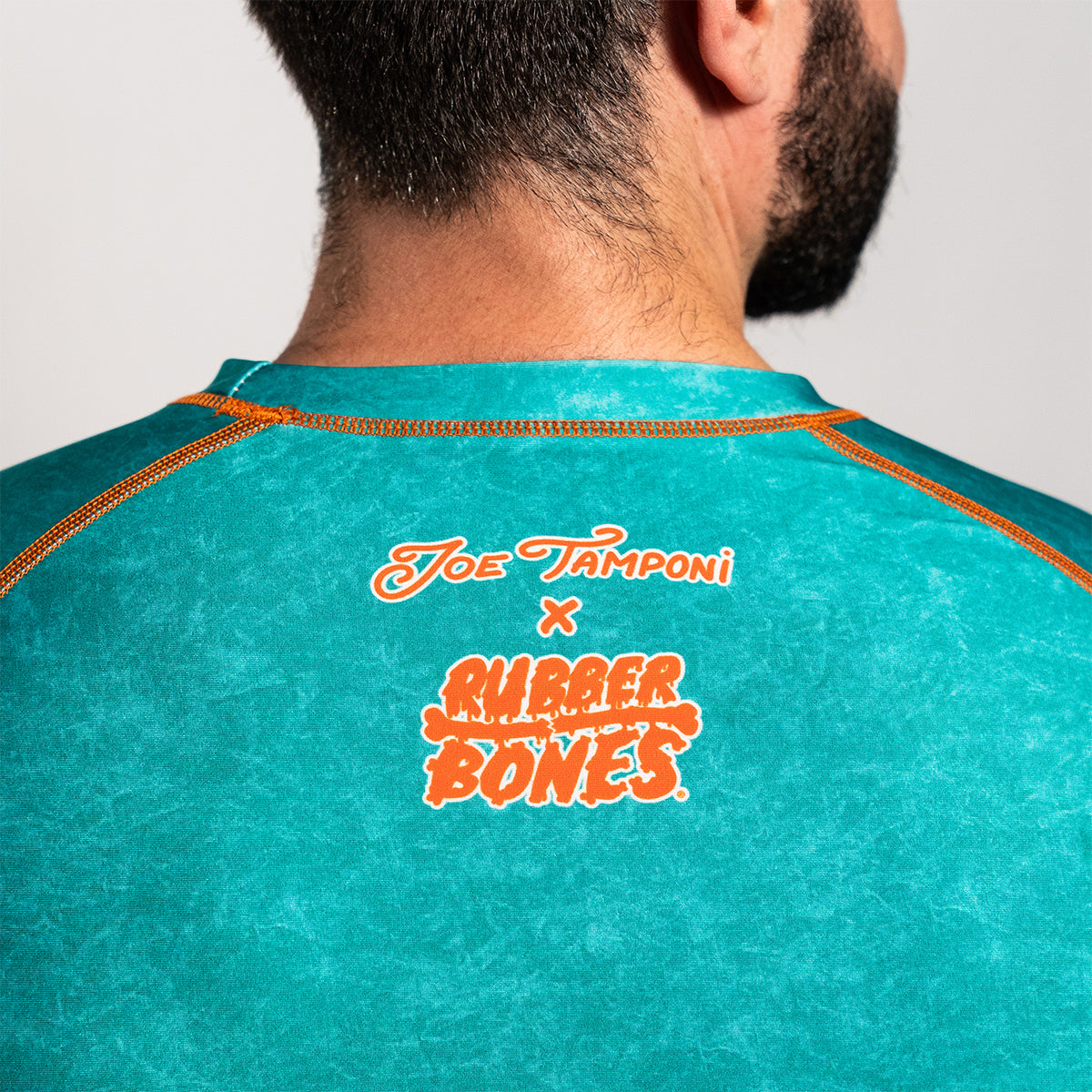 Back view of the rash guard, showing “Joe Tamponi x Rubber Bones” printed in orange and white lettering at the upper center below the collar, with contrast orange stitching.
