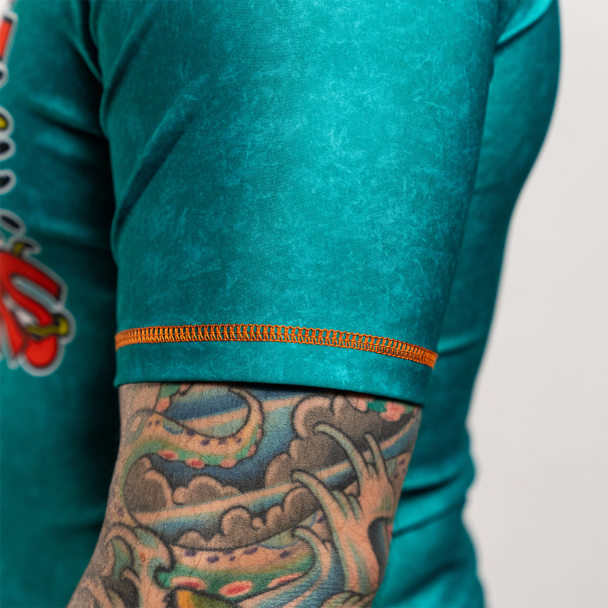 Close-up of the opposite sleeve of the same teal rash guard, showing orange stitching detail and a tattooed forearm. This sleeve does not have a patch or logo.