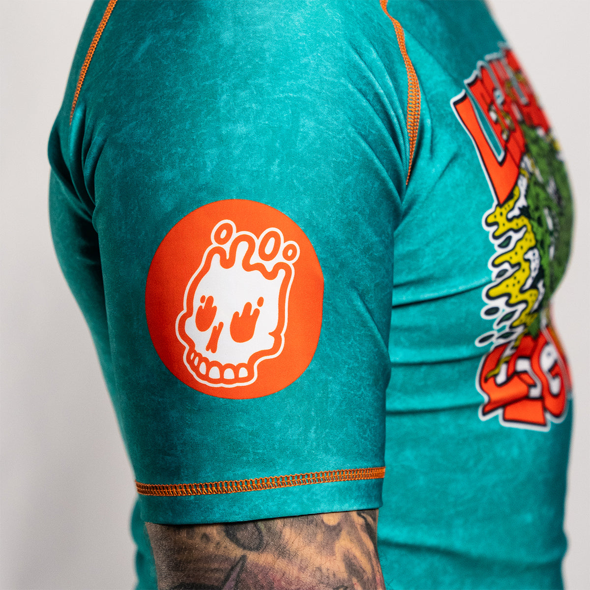 Close-up of a teal short-sleeve rash guard with orange stitching. The upper sleeve features a circular orange patch with tje Rubber Bones white skull icon.