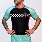A person wearing a short-sleeve MintDrift rash guard with teal sleeves, black torso, and pink side panels. The chest features a white checker stripe and skull logo on the left side.