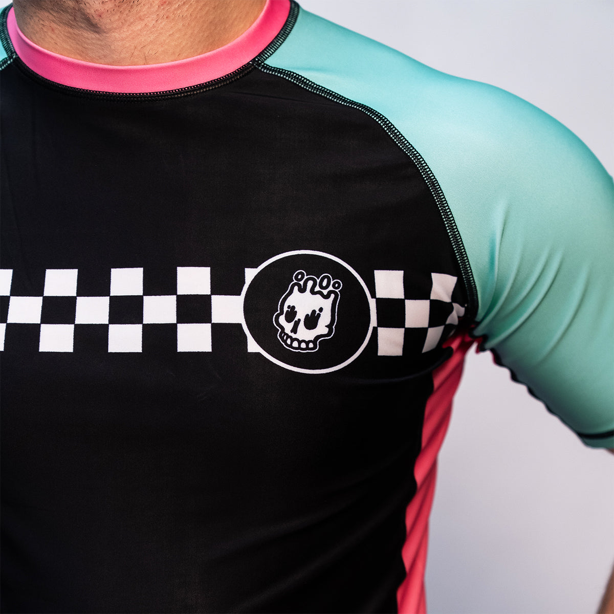 Close-up view of the chest area on the rash guard, showing the black torso with a white checker stripe and skull logo on the left side. Teal sleeve and pink collar also visible.