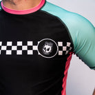 Close-up view of the chest area on the rash guard, showing the black torso with a white checker stripe and skull logo on the left side. Teal sleeve and pink collar also visible.