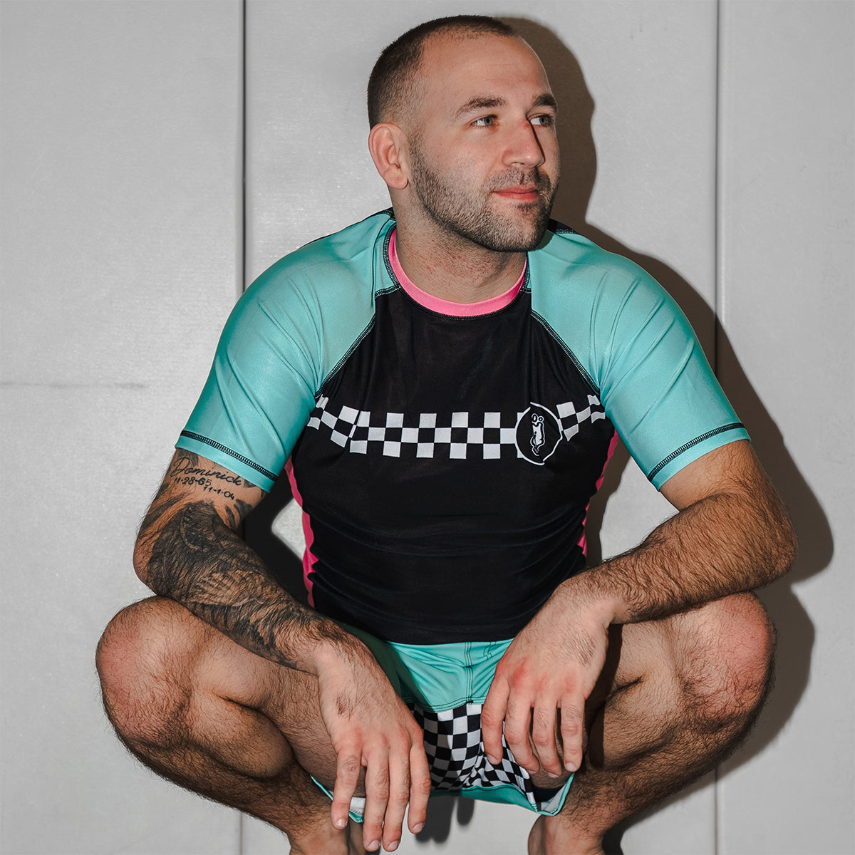 A person squatting against a white wall while wearing the Mint Drift rash guard and matching shorts. The checker pattern runs across the chest and shorts crotch panel.