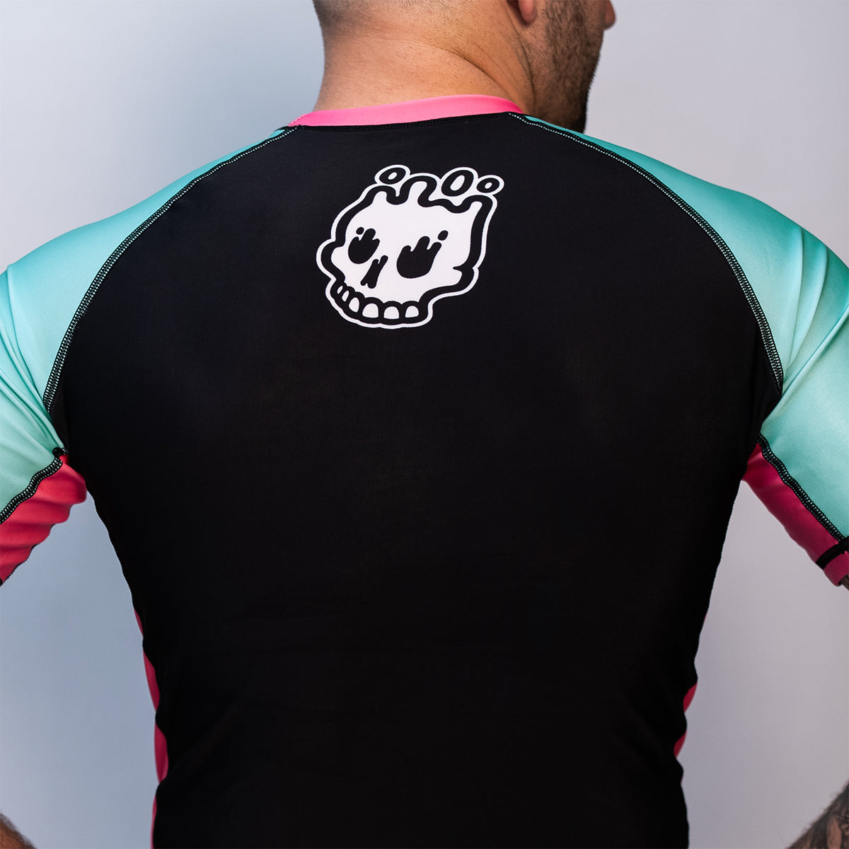 Back view of the rash guard worn by a person. The back is solid black with teal sleeves and pink side panels. A white skull logo is printed at the upper center back.