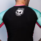 Back view of the rash guard worn by a person. The back is solid black with teal sleeves and pink side panels. A white skull logo is printed at the upper center back.