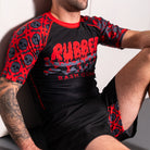 Close-up of a seated model wearing a black and red BJJ rash guard. The focus is on the torso, highlighting the dripping red logo and red skull graphics on the short sleeves. The black and red BJJ shorts are partially visible.