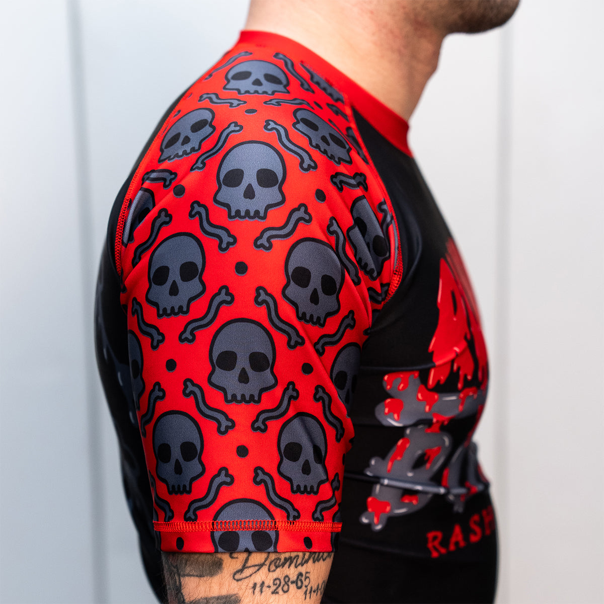 Side view of a men's black and red short-sleeve BJJ rash guard. Close-up of the right sleeve, showing a red background with a repeating pattern of black skulls and bones. The model's tattooed arm is partially visible.