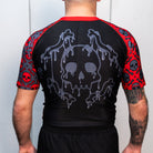 Back view of a men's black and red short-sleeve BJJ rash guard. The rash guard has a large melting skull graphic in gray on the back, with red skull and bone graphics on the sleeves. The model is standing with their back to the camera.