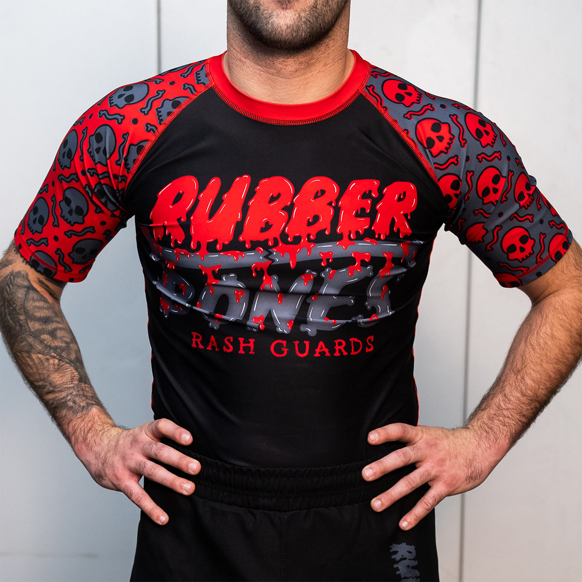 Front view of a men's black and red short-sleeve BJJ rash guard. The rash guard features a dripping red and gray logo on the chest, with red skull and bone graphics on the sleeves. The model stands with hands on hips.