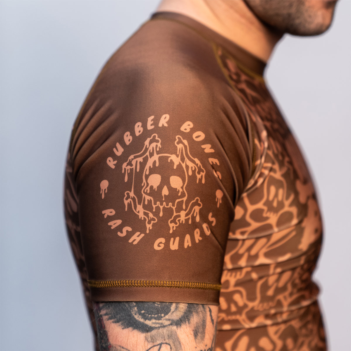 Close-up of the Rubber Bones Ranked Brown rash guard sleeve – Melting skull logo with 'Rubber Bones Rash Guards' text on a brown background.