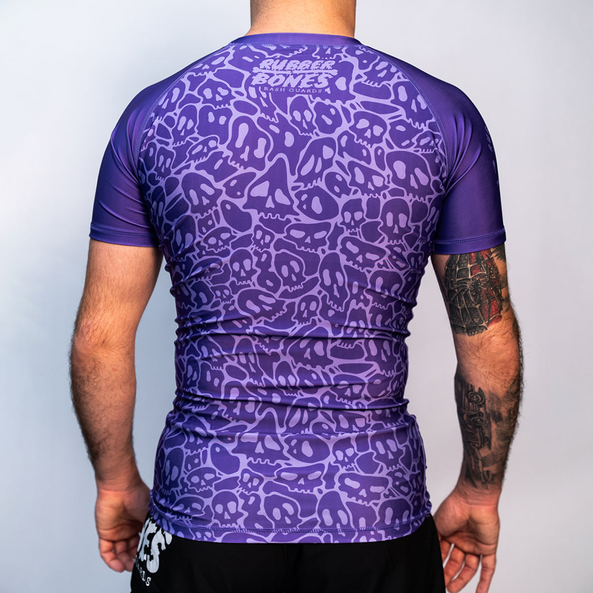 Back view of the Rubber Bones Ranked Purple BJJ rash guard – Purple short-sleeve rash guard with an all-over skull pattern and Rubber Bones branding on the upper back.