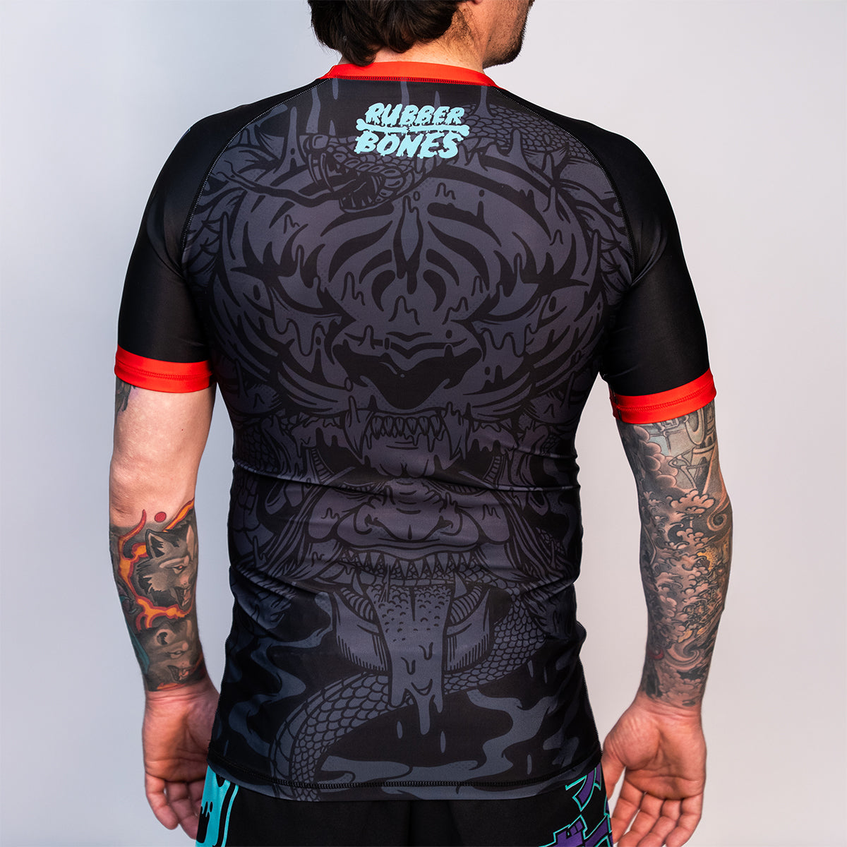 Back view of the Rubber Bones Tora Oni men's rash guard – Black rash guard with a tonal tiger-demon design, Rubber Bones branding, and red sleeve cuffs.