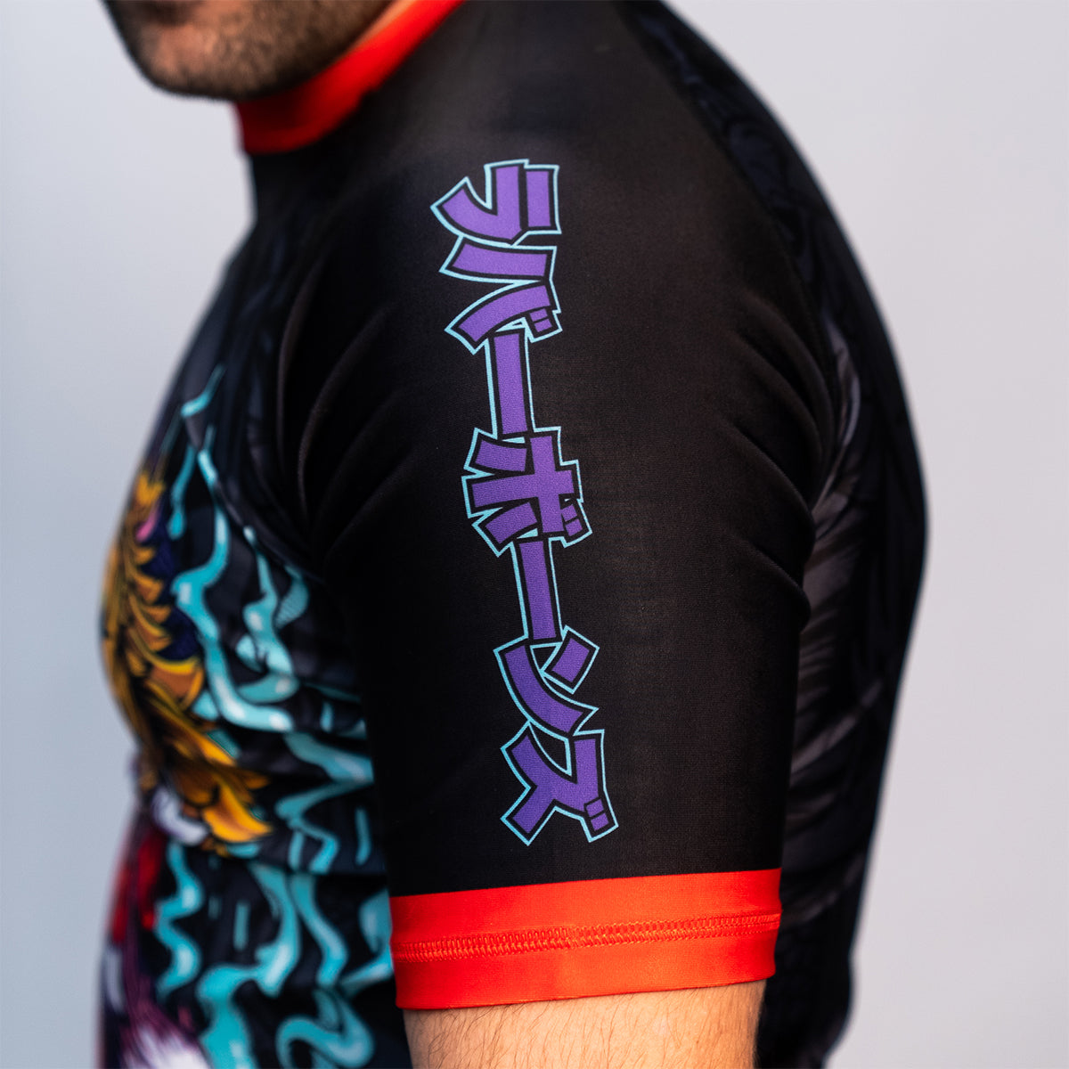 Side view of the Rubber Bones Tora Oni men's rash guard sleeve – Japanese lettering in purple outlined in teal on a black background with red stitching.