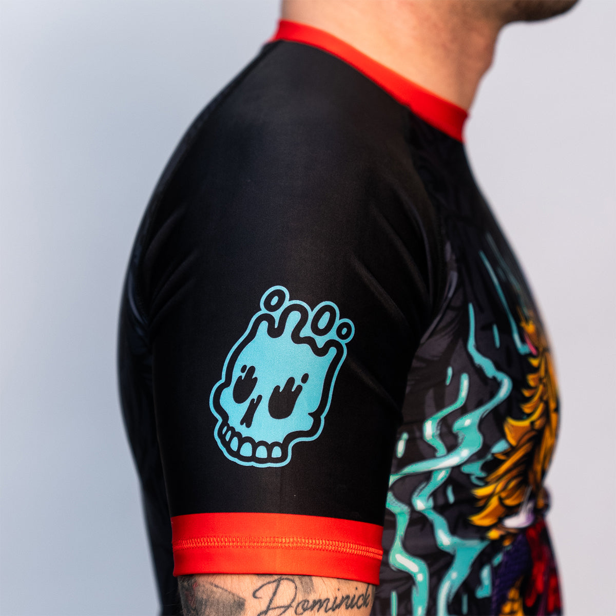 Close-up of the Rubber Bones Tora Oni men's rash guard sleeve – Teal melting skull graphic on a black background with red stitching.