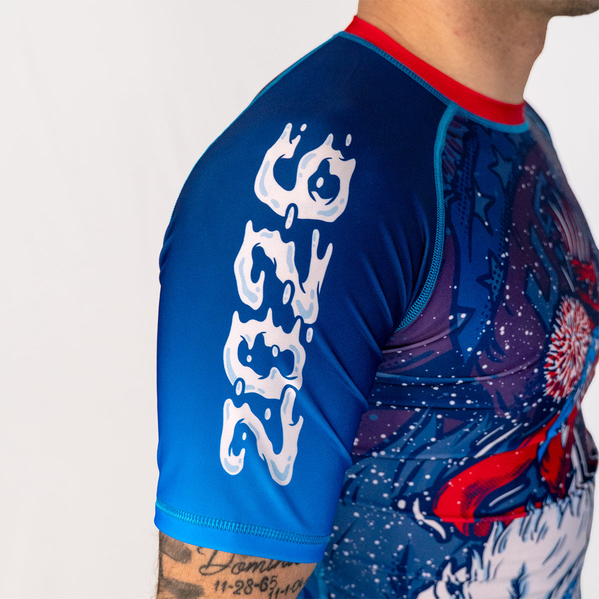 Side profile of Men's Yeti Games rash guard showing athletic compression fit, red neckline, blue sleeves, and detailed skeleton skier artwork on torso.