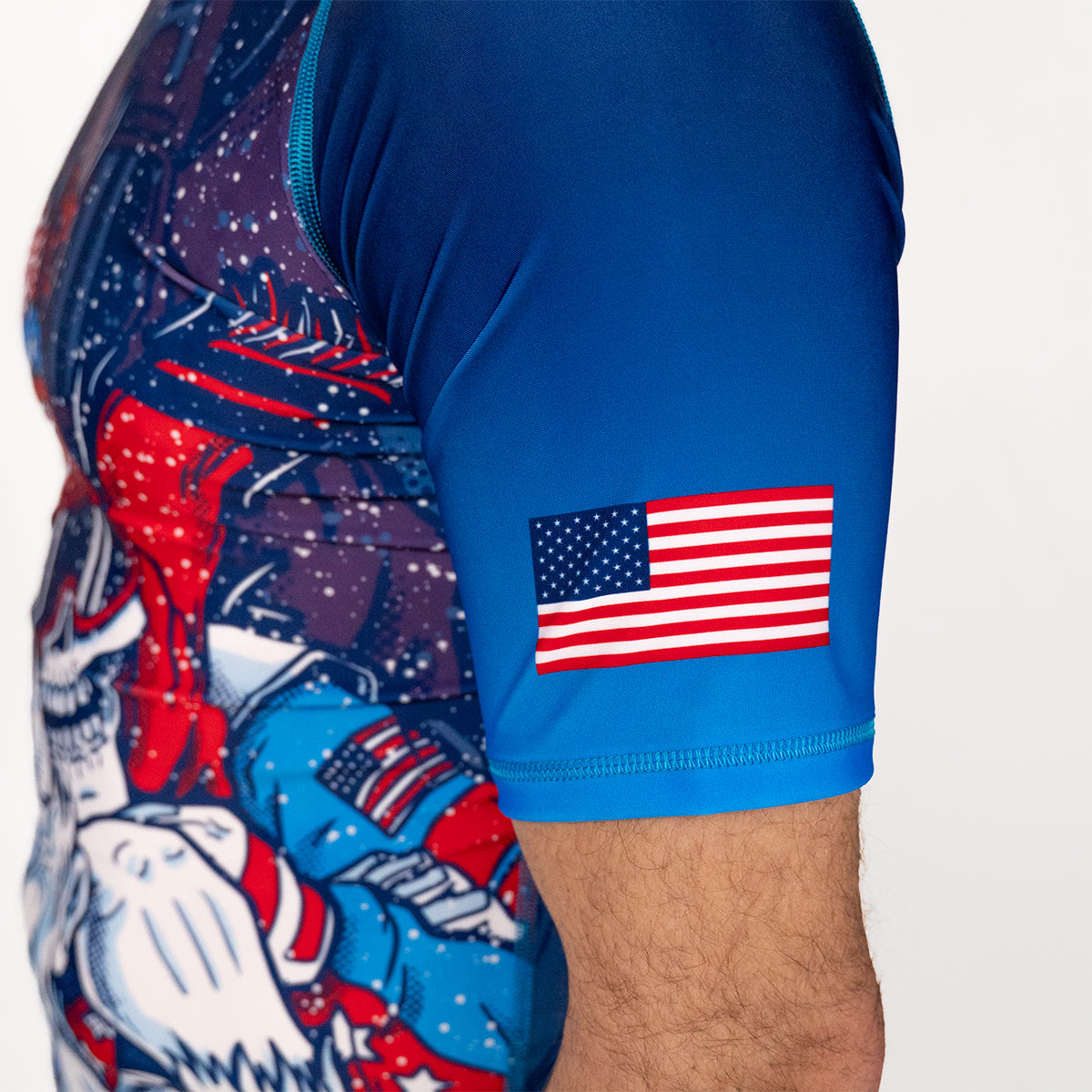 Close-up of Men's Yeti Games rash guard sleeve showing USA flag patch, skeleton skier Yeti artwork in red and blue, and blue compression sleeve construction.