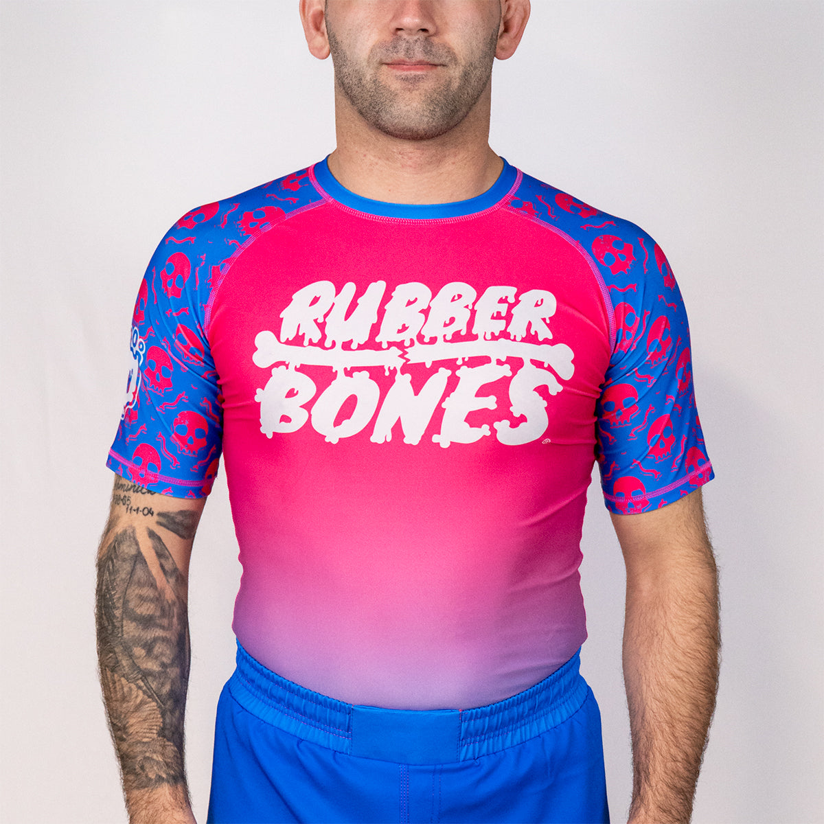 Front view of the Rubber Bones Toxic Berry men’s no gi BJJ rash guard from the Deadly Harvest Collection featuring pink body and blue skull print sleeves.